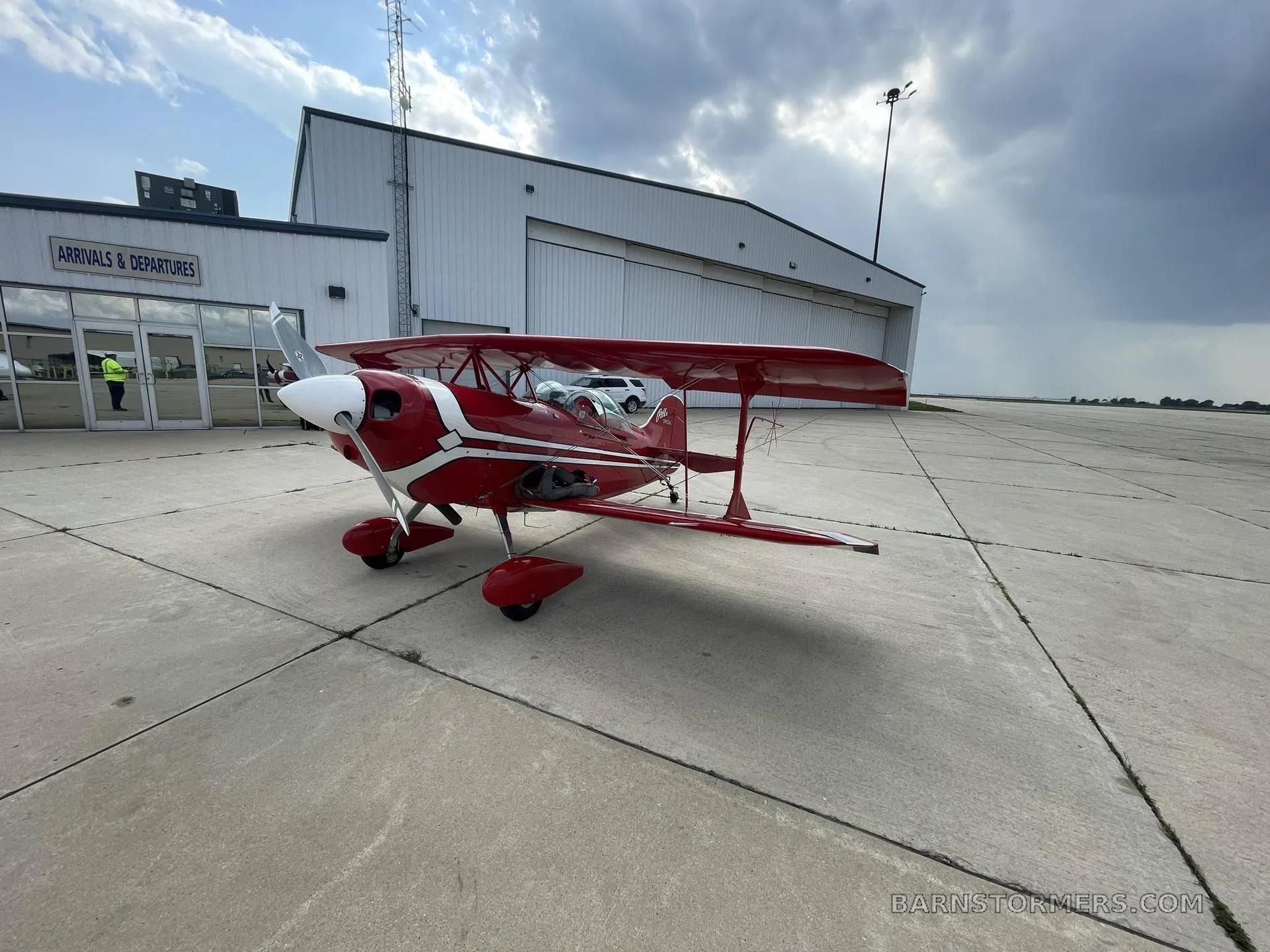 A red and white Pitts Special biplane is parked on an airfield tarmac next to a white hangar under a cloudy sky.