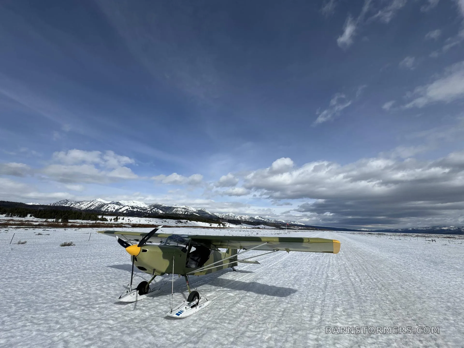 A camouflage-painted light aircraft with skis is parked on a snowy field with mountains in the background under a partly cloudy sky.