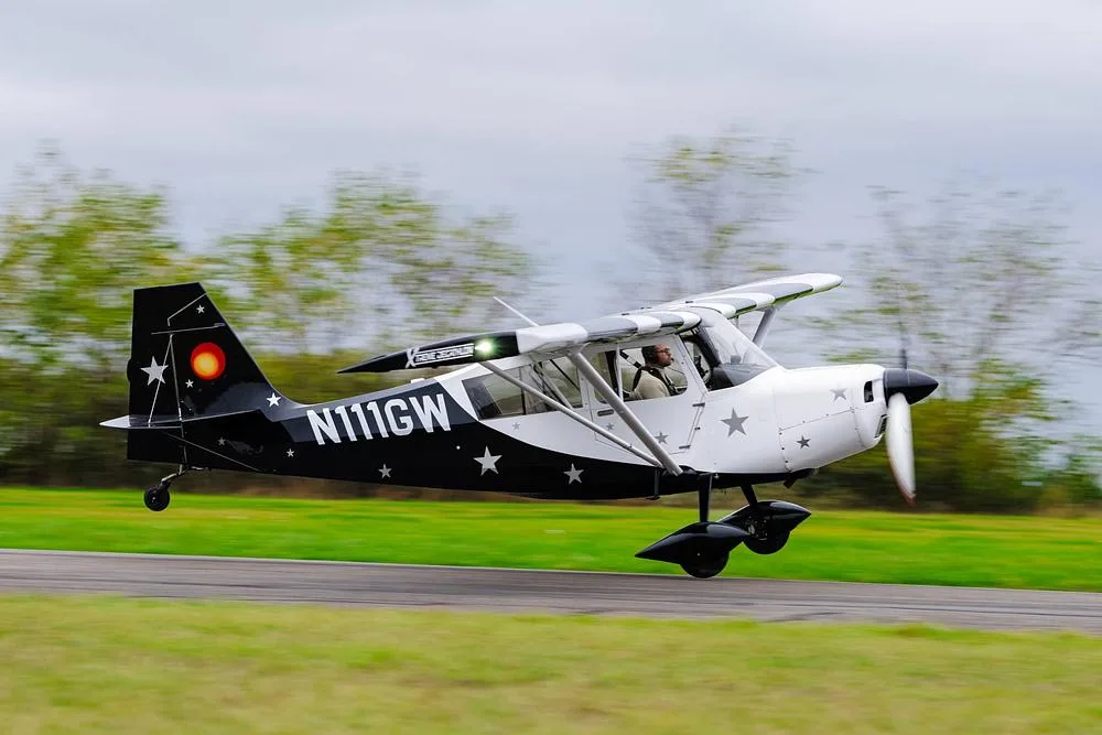 A white and black high-wing monoplane with star decals, registered N111GW, taking off from a grassy runway.