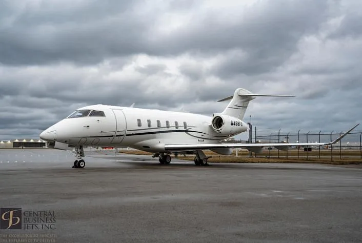 A white Bombardier Challenger business jet with dark accents is parked on a wet tarmac under a cloudy sky.