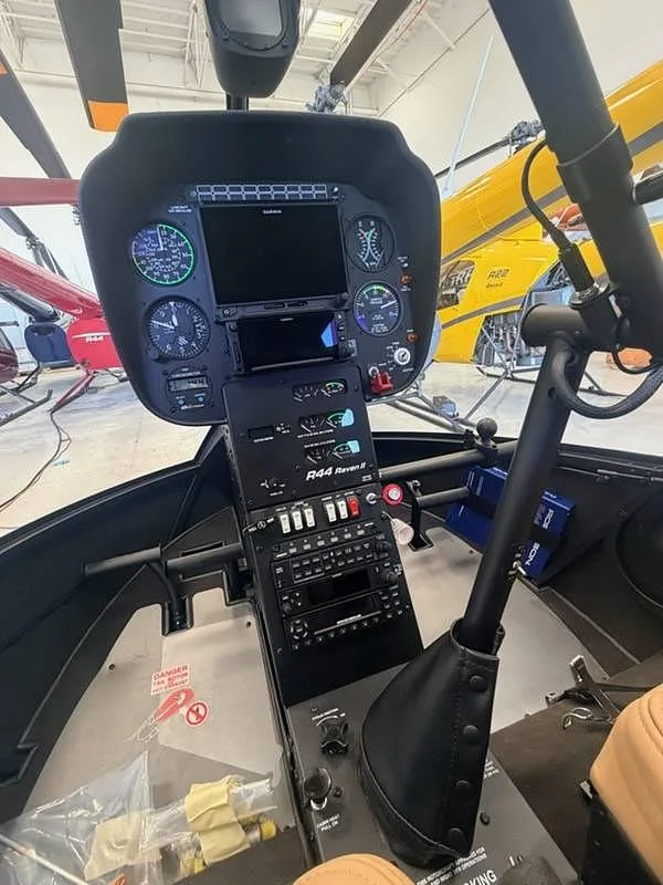 The cockpit instrument panel and controls of a Robinson R44 Raven II helicopter, with other helicopters in the background.