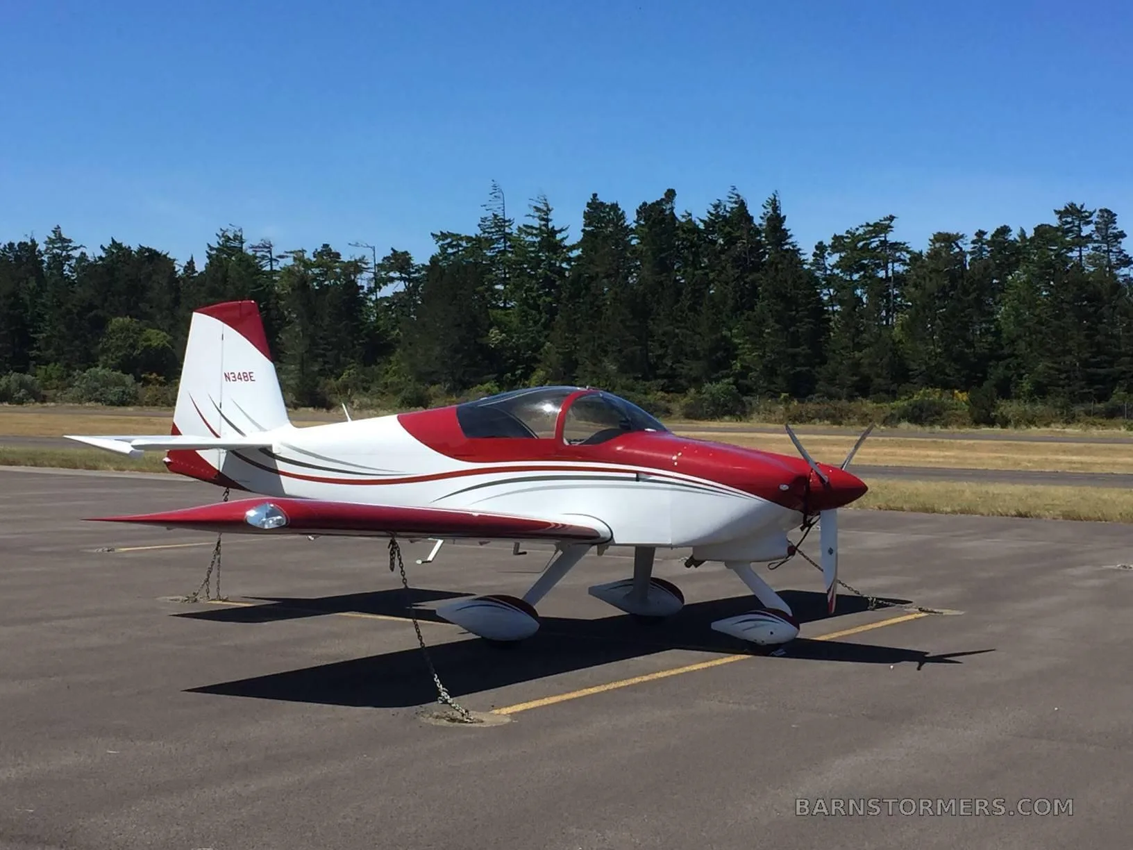 A red and white single-engine aircraft (N34BE) is parked on an asphalt runway with trees in the background under a clear blue sky.