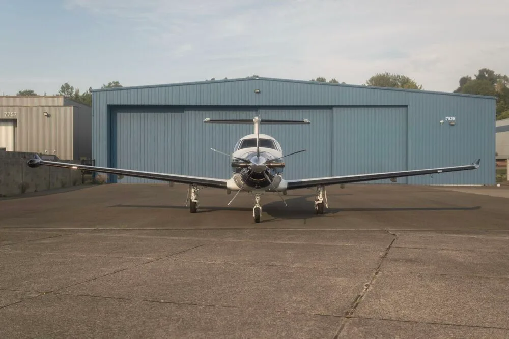 A white and blue Pilatus PC-12 aircraft with registration N226N seen from a head-on view, parked in front of a blue hangar.