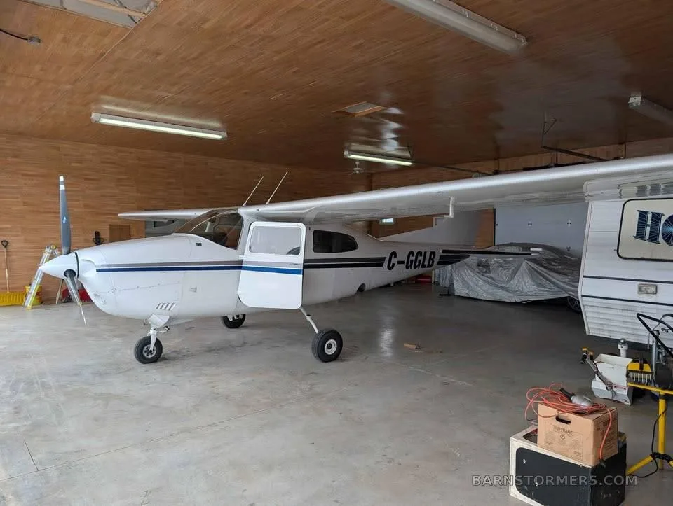 A white Cessna 182 with blue stripes, registration C-GGLB, parked in a hangar with its door open.