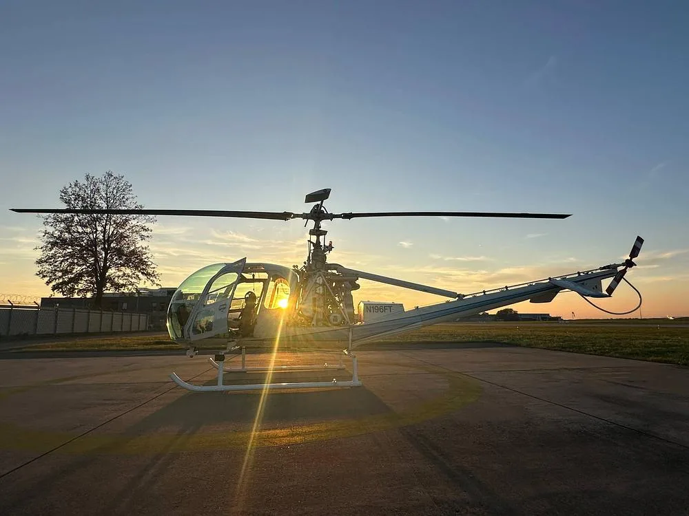 A white Hiller UH-12 helicopter with registration N196FT sits on an airfield at sunset.