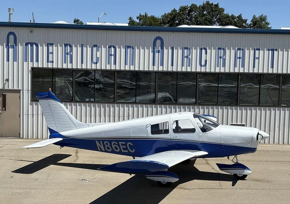 A white and blue Piper Cherokee aircraft (N86EC) parked on the tarmac in front of a hangar.