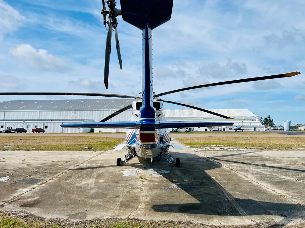 Rear view of a white and blue helicopter with its tail rotor and horizontal stabilizer visible, parked on an airfield.