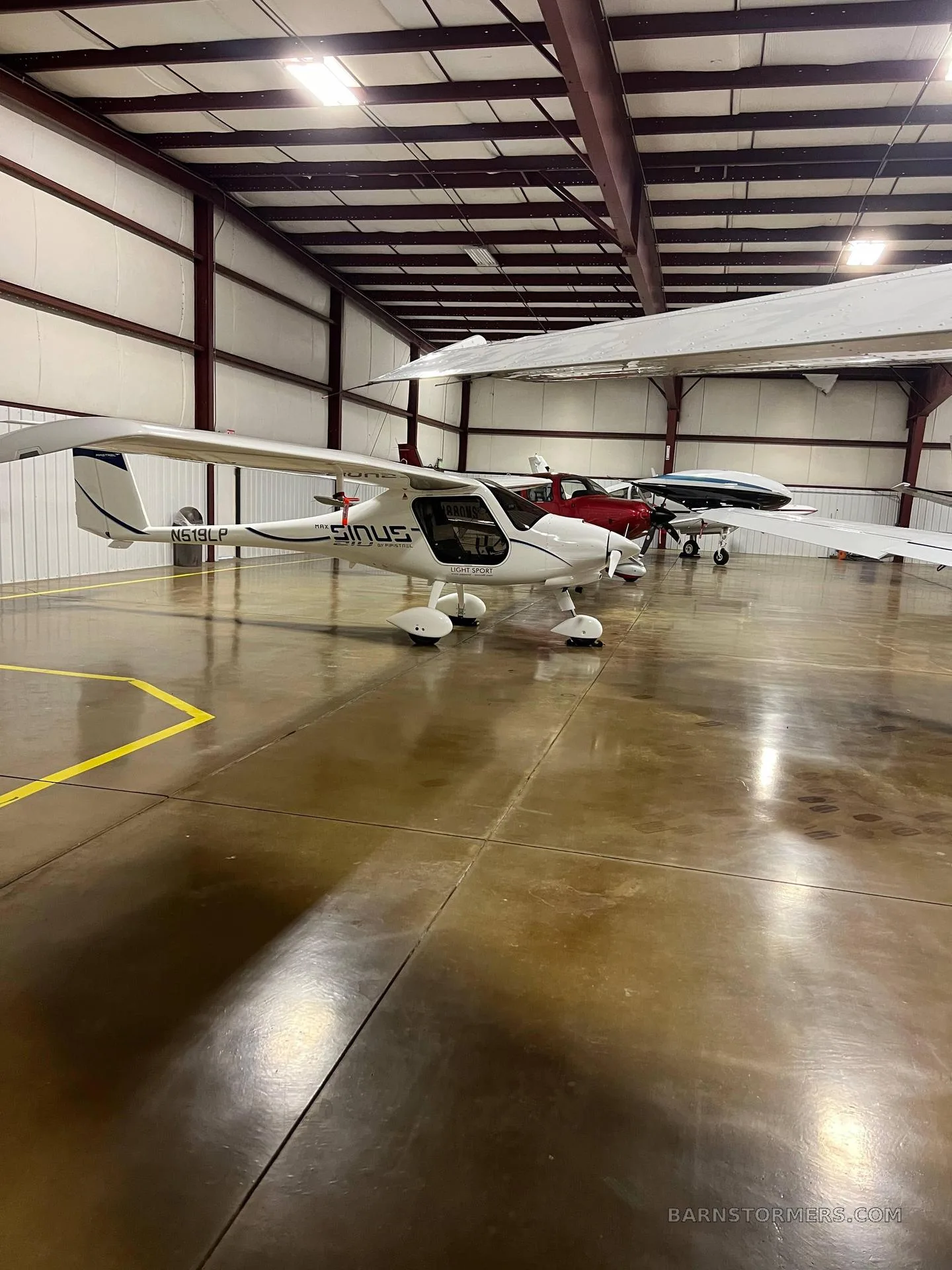 A white Pipistrel Sinus light sport aircraft with registration N618LP is parked in a hangar, with other aircraft visible in the background.
