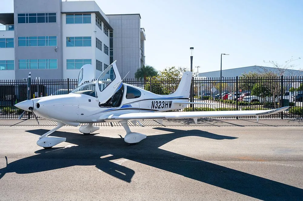 A white Cirrus SR22 aircraft with blue and black stripes, registration N323HW, parked with its gull-wing doors open on an asphalt ramp.