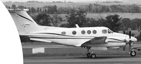 A white twin-engine turboprop aircraft with dark stripes on its fuselage, seen from the side on an airfield.