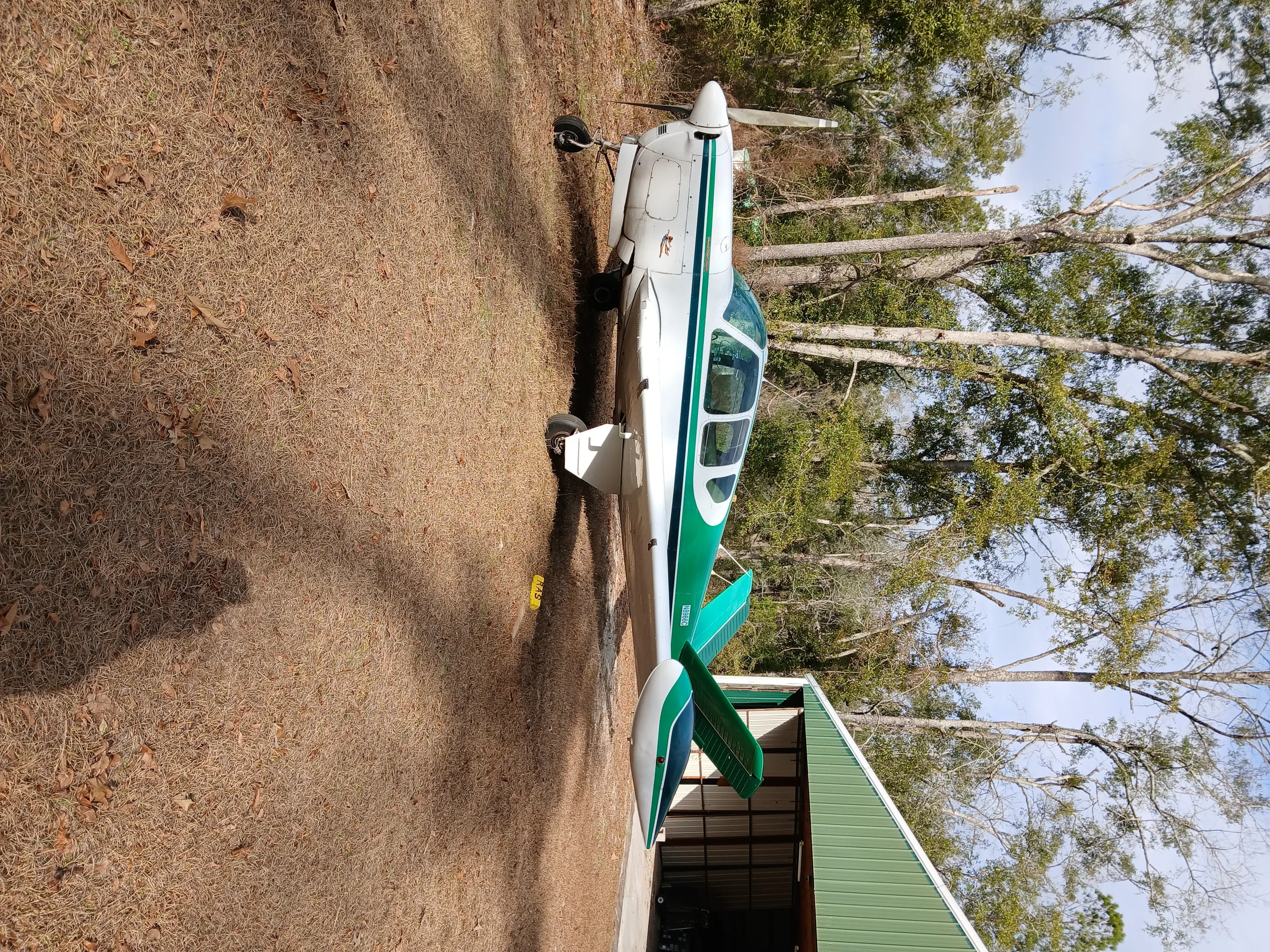 A white and green single-engine aircraft is parked on a grassy area next to a green hangar.