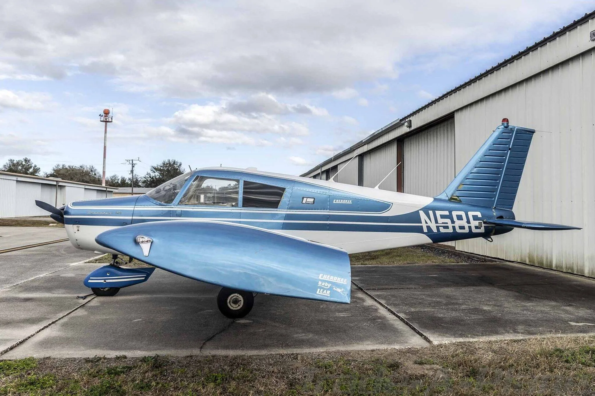 A blue and white Piper Cherokee N586 parked on an asphalt ramp next to a hangar.