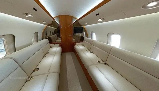 The spacious interior of a private jet cabin featuring two long, opposing beige leather sofas and wooden paneling.