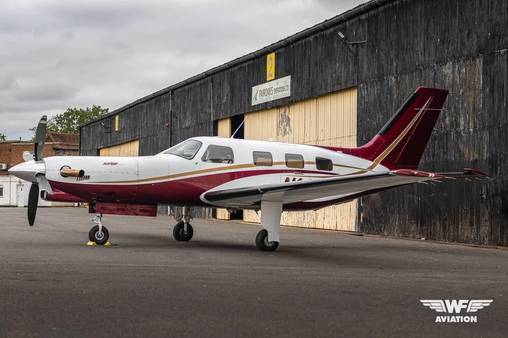A white and maroon single-engine turboprop aircraft with gold trim parked on a tarmac in front of a dark hangar.