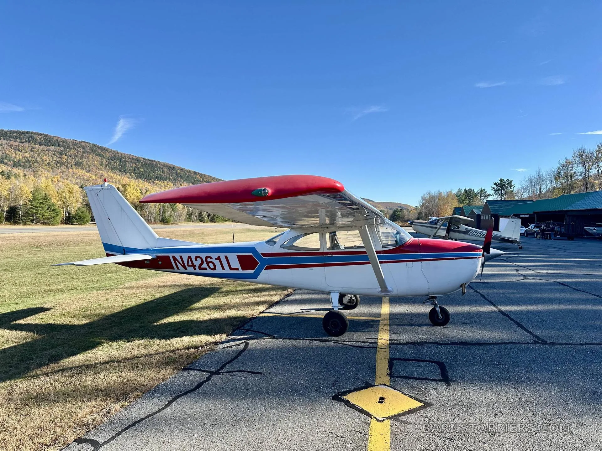 A white single-engine propeller aircraft with red and blue stripes, viewed from the rear-left, parked on an asphalt ramp with other aircraft and hangars in the distance.