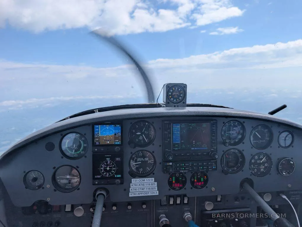 A view from the cockpit of a small aircraft, showing the instrument panel, controls, and a propeller blur against a cloudy sky.