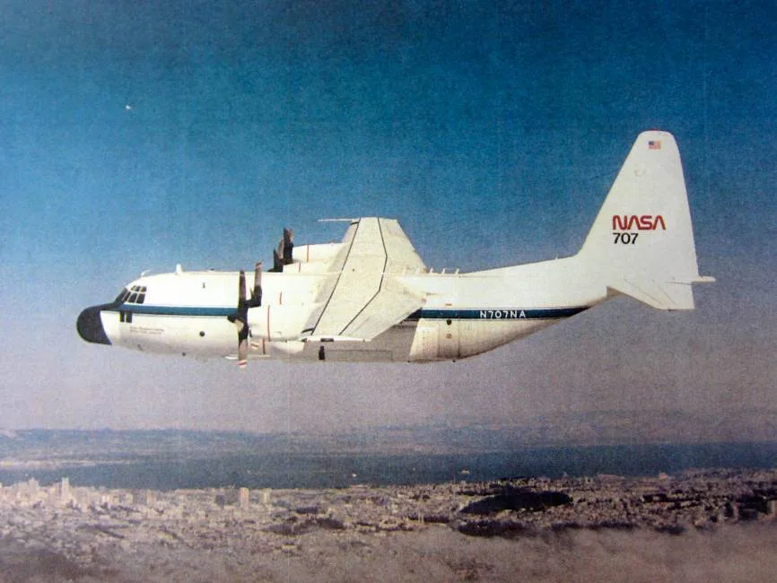 A white NASA C-130 aircraft with blue stripes flies over a city.