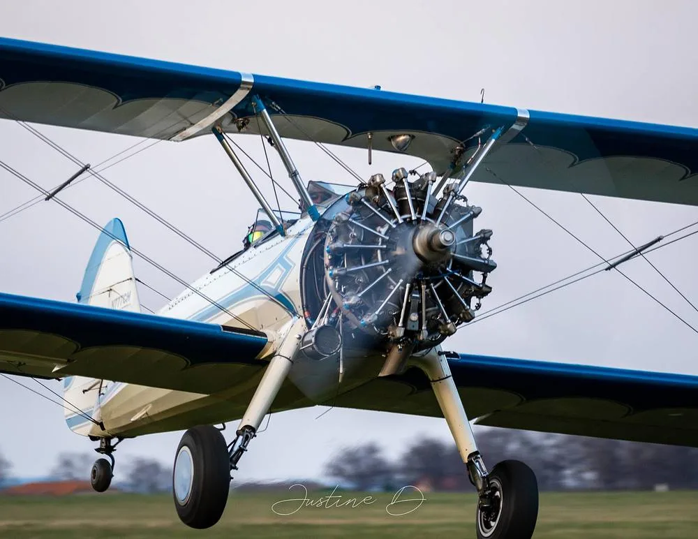A white and blue biplane, N777CH, with a radial engine, flying low over a grassy field.