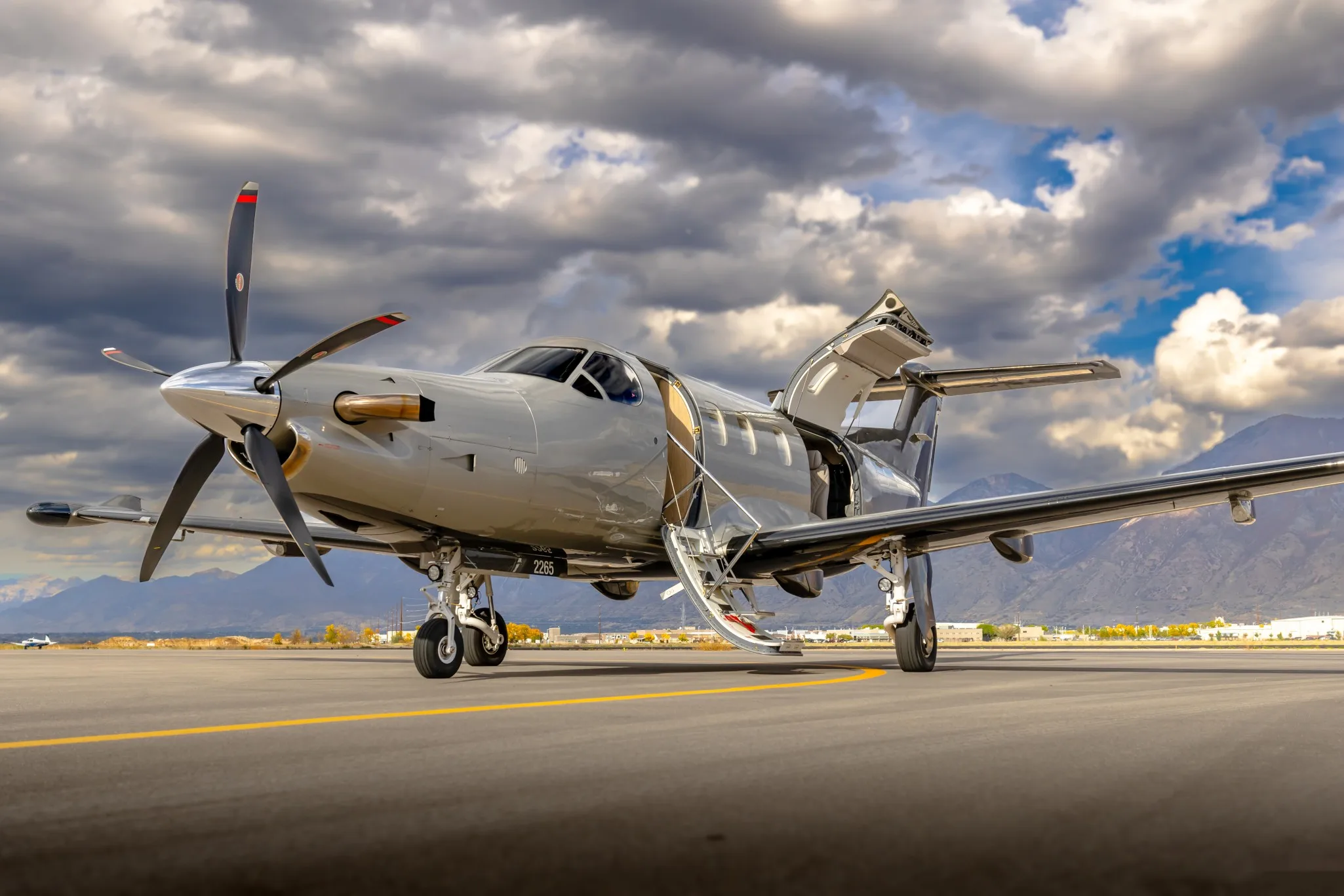 A silver Pilatus PC-12 turboprop aircraft with its main door open is parked on an airport tarmac with mountains in the background.
