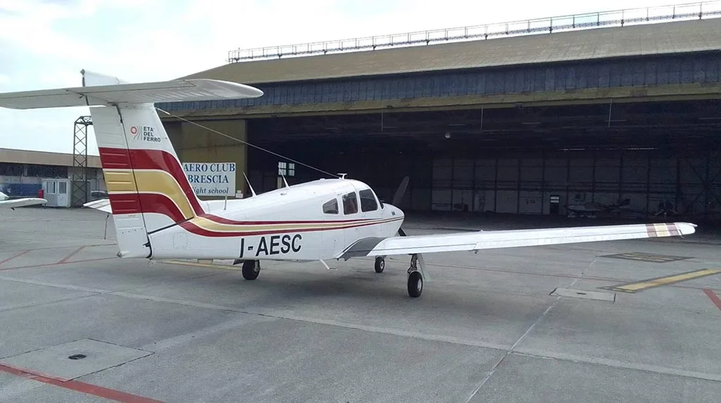 A white Piper PA-28 aircraft with red and gold stripes parked on an airfield in front of a large hangar.