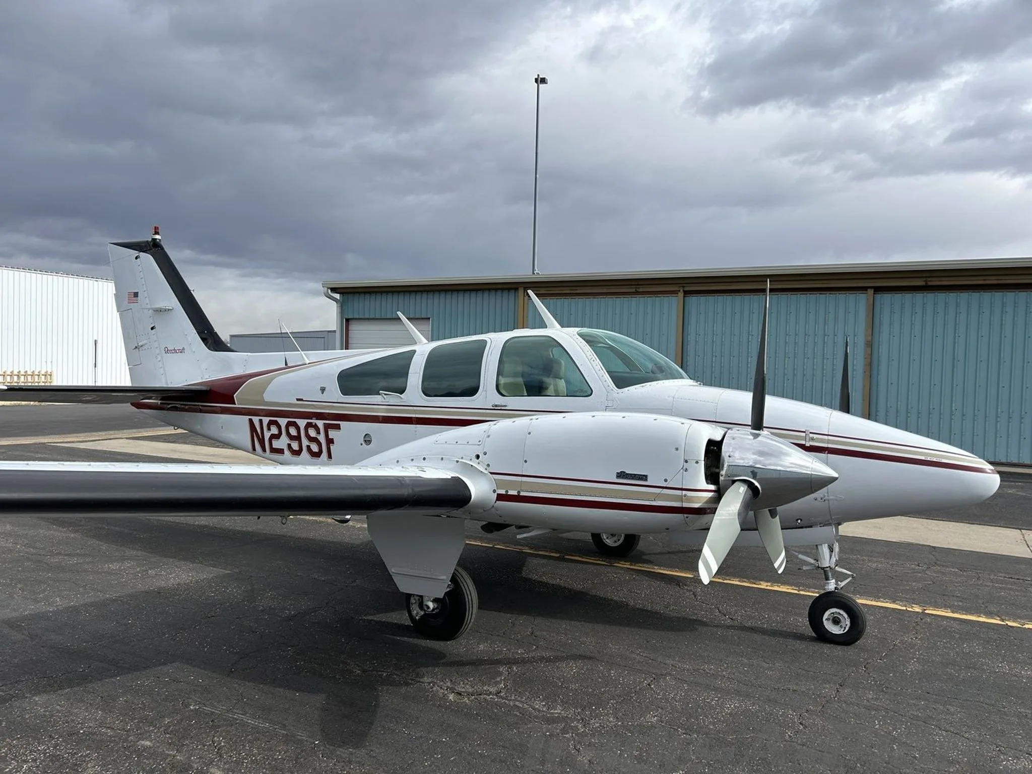 A white and maroon Beechcraft Baron N29SF parked on an asphalt ramp with hangars in the background under a cloudy sky.