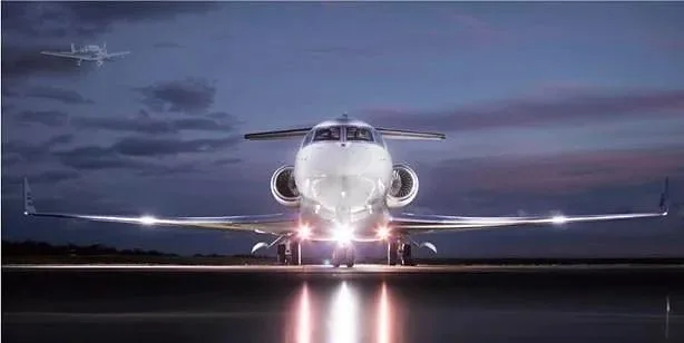 A white business jet with its landing lights on, parked on a tarmac at dusk, with another aircraft flying in the distant sky.