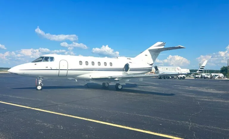 A white business jet with dark blue stripes on the fuselage and tail, parked on an asphalt tarmac under a blue sky.