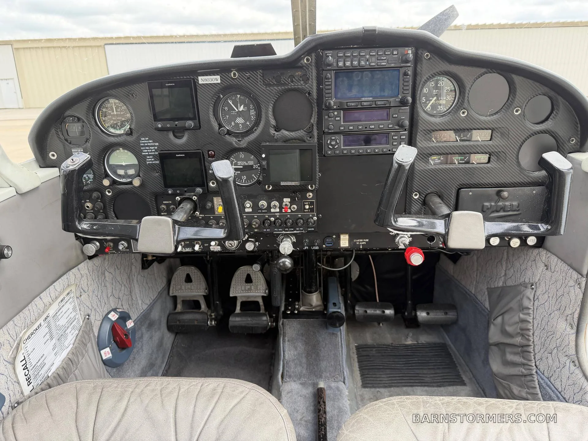 The cockpit of a Piper Cherokee, showing the instrument panel with various gauges and screens, yokes, and pedals.
