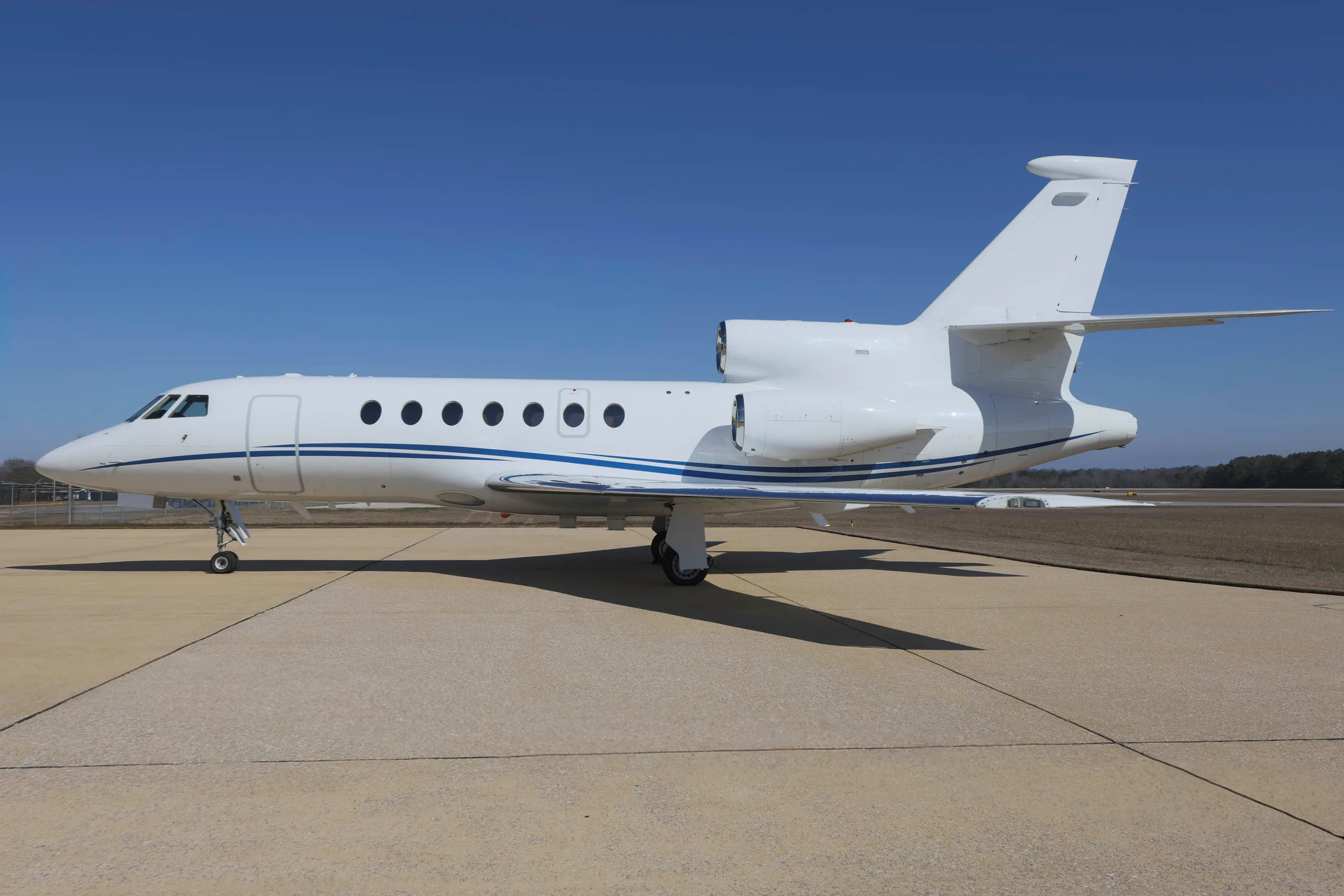 A white business jet with a blue stripe is parked on a tarmac under a clear blue sky.