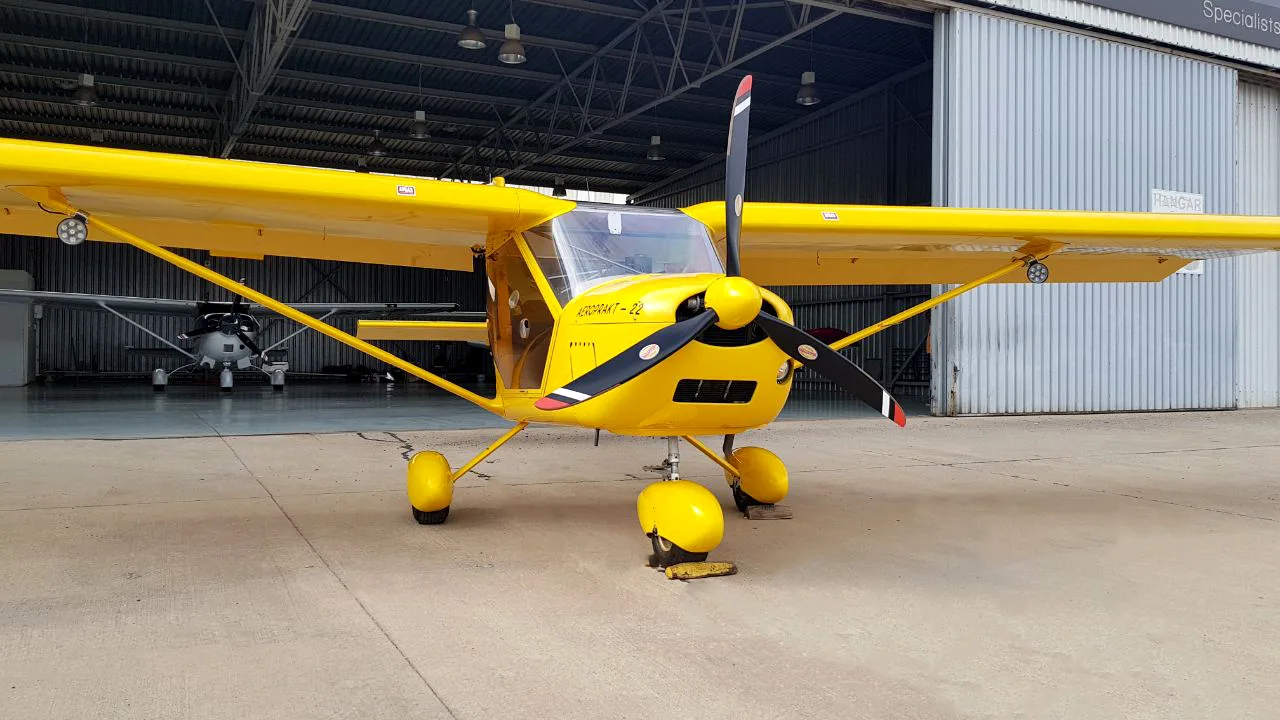 A bright yellow Aeroprakt-22 aircraft is parked on concrete outside an open hangar.