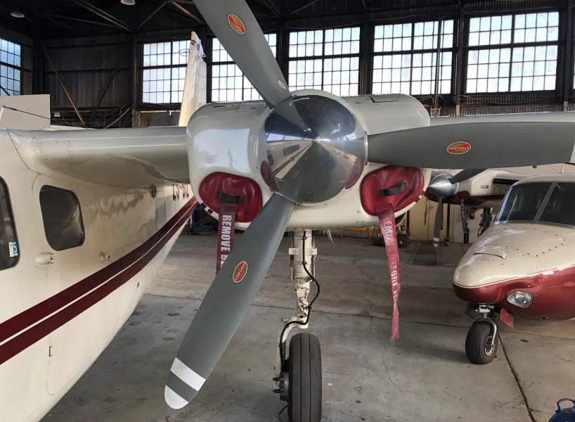 Front view of the right engine and propeller of a white aircraft with a maroon stripe, inside a hangar, with another aircraft nose visible.