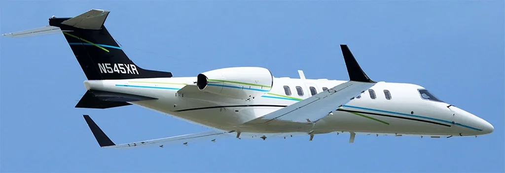 A white business jet with blue and green stripes and registration N545XR on its black tail, flying against a clear blue sky.