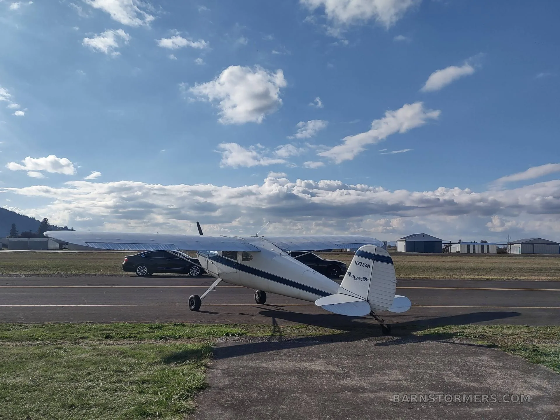 A white and blue taildragger aircraft is parked on a tarmac, seen from the rear-right, with a car and hangars in the background under a partly cloudy sky.