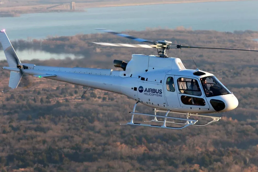 A white Airbus Helicopters H125 (AS350) flies over a wooded landscape with a body of water in the background.