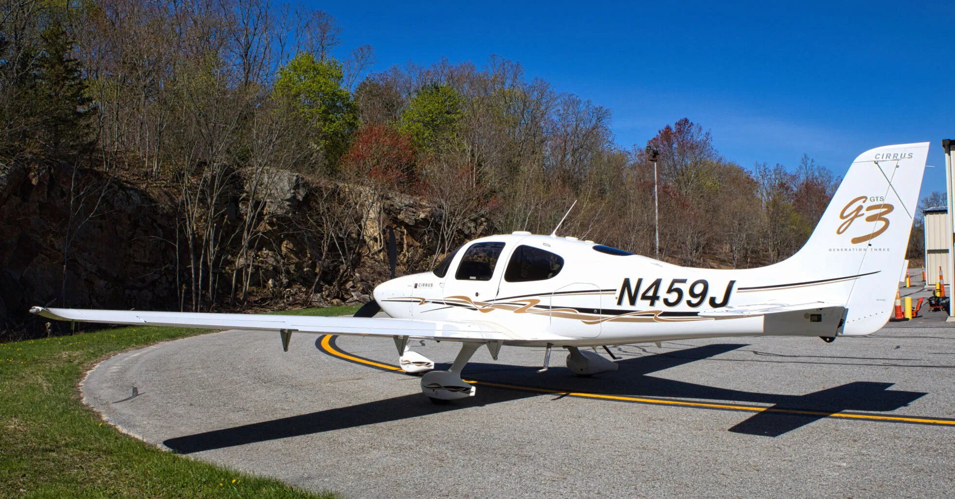 A side view of the white Cirrus SR22 GTS Generation Three aircraft (N459J) with gold and black stripes, parked on an airfield.