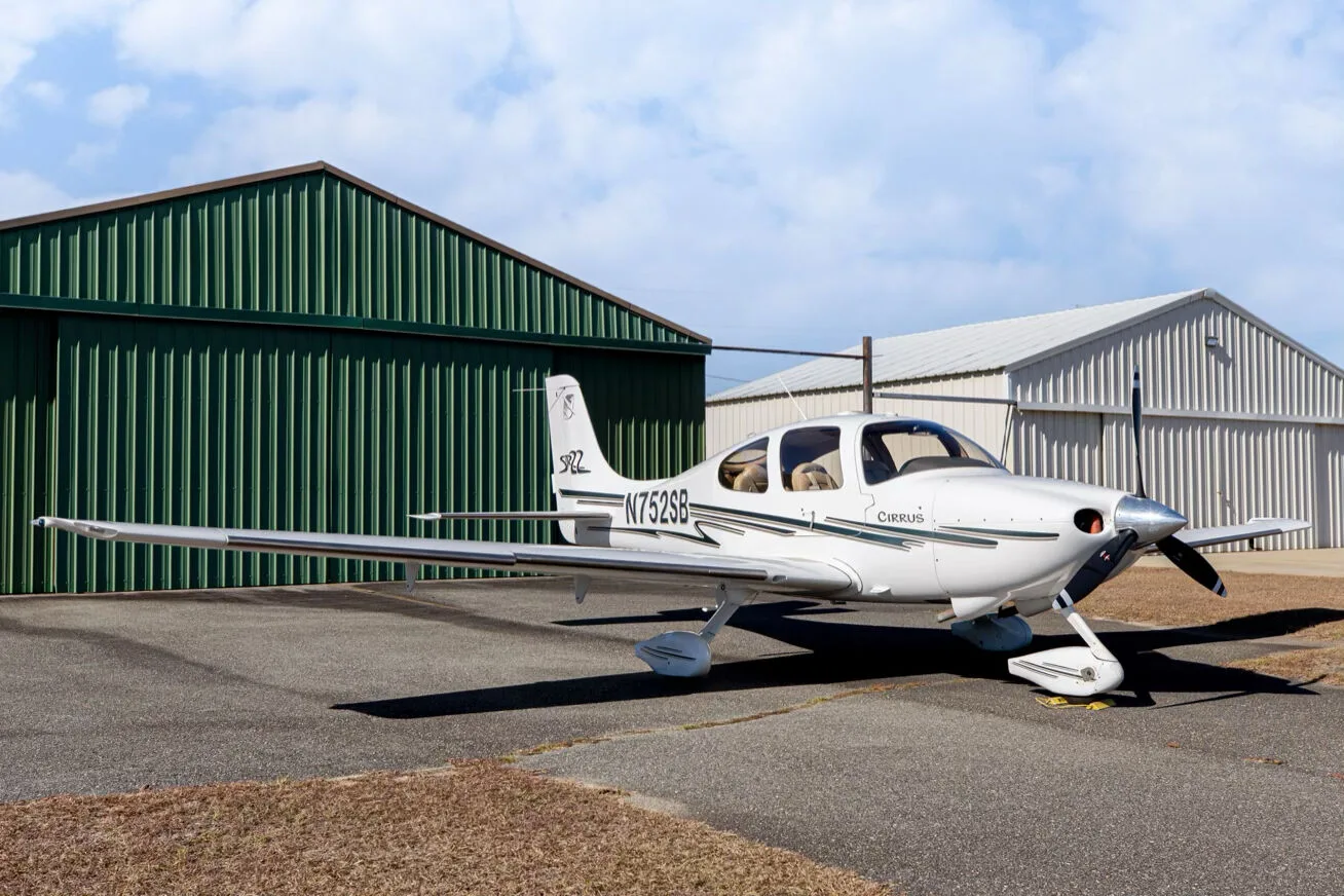 A white Cirrus SR22 aircraft with green and grey stripes, registration N752SB, parked on an asphalt ramp next to a green hangar under a partly cloudy sky.