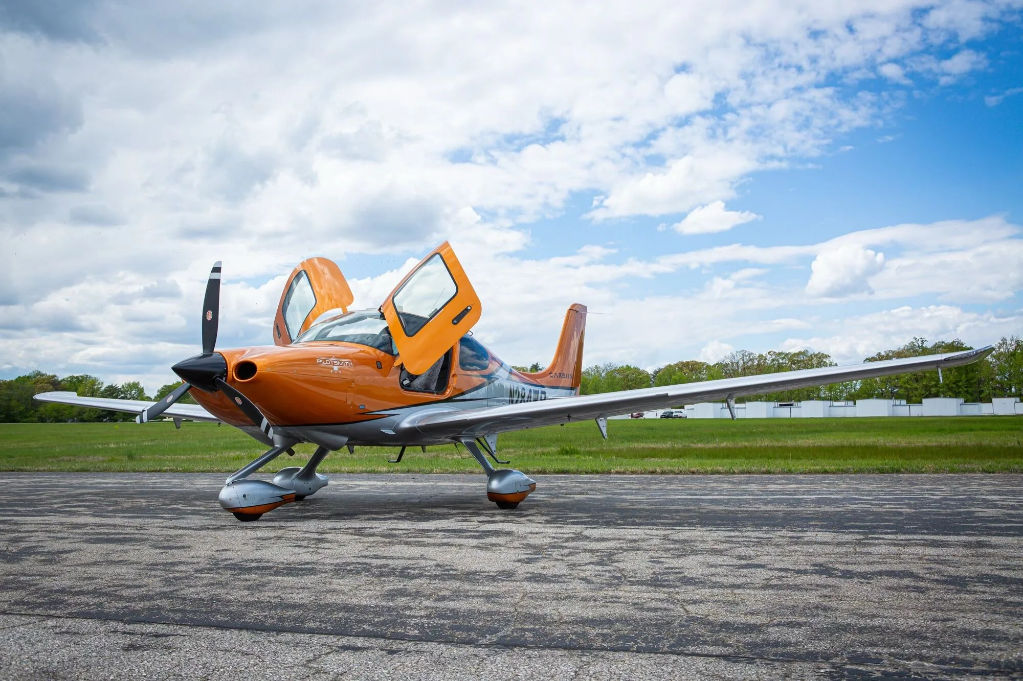 An angled front view of an orange and silver Cirrus SR20 aircraft, N284WB, with its gull-wing doors open on an airfield.
