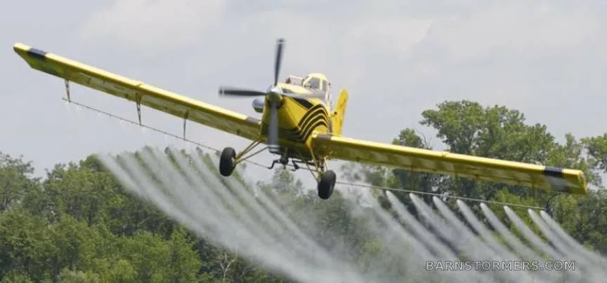 A yellow agricultural aircraft with black stripes is flying low over trees, actively spraying liquid.
