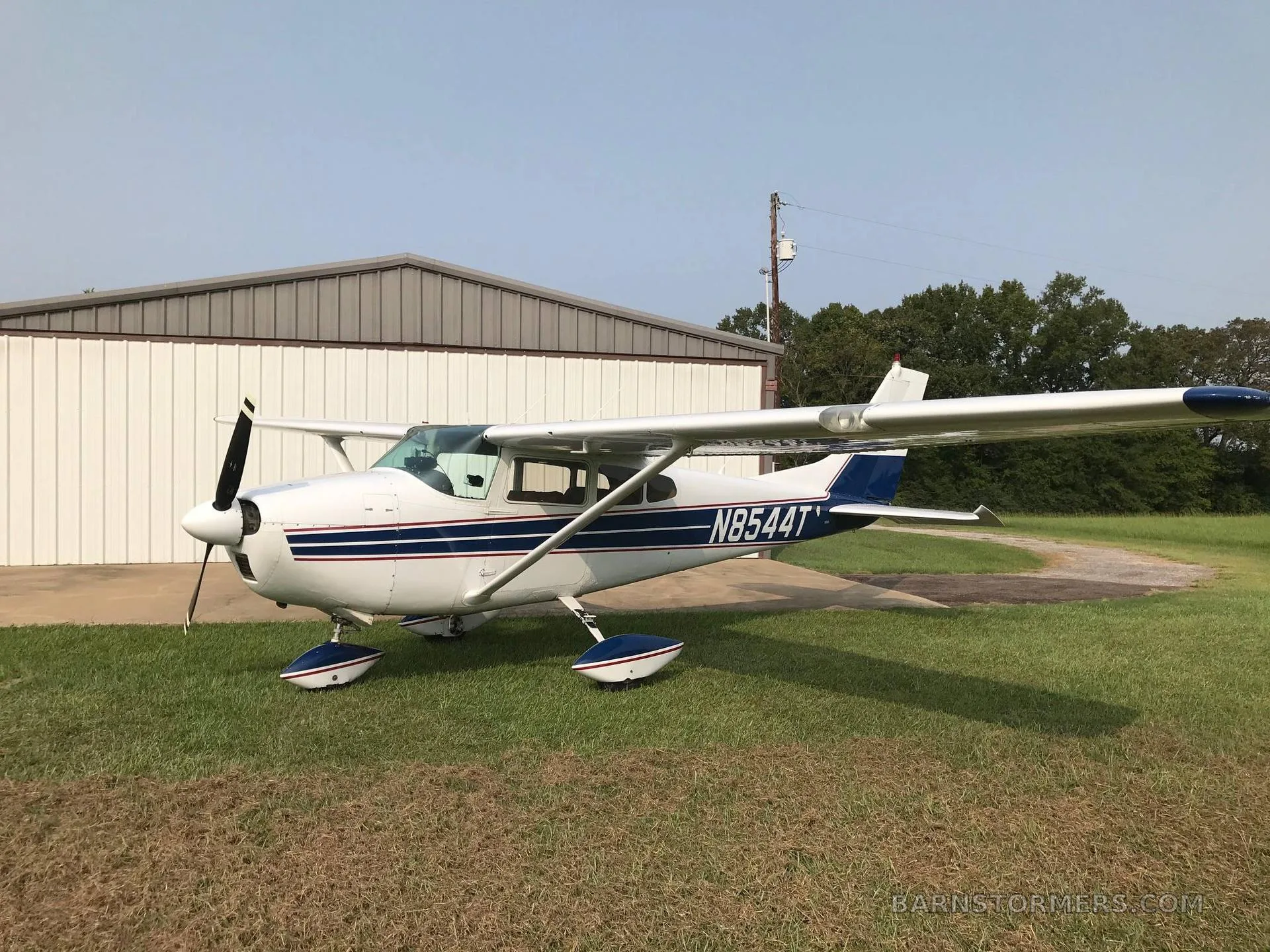 A white and blue Cessna 182 aircraft is parked on grass next to a hangar.