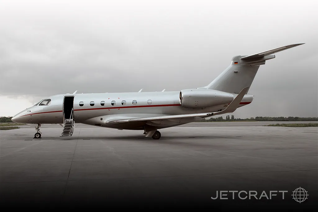 A silver Embraer Legacy business jet with a red stripe, door open, parked on an airfield under a cloudy sky.