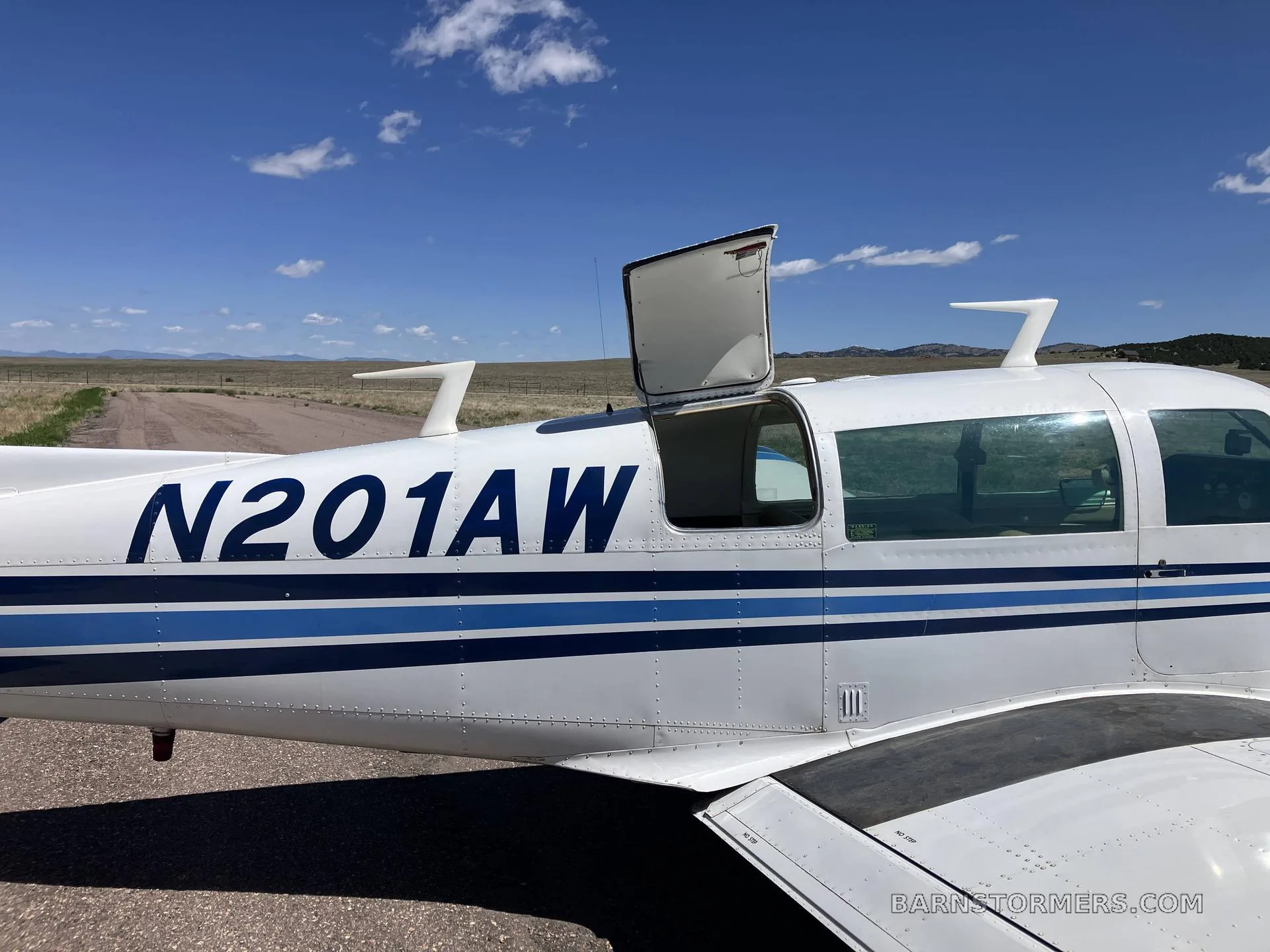 A close-up side view of a white Mooney M20J aircraft, registration N201AW, with its baggage door open.