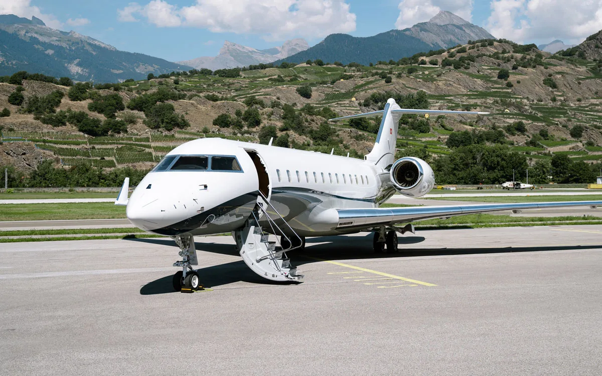 A white and dark grey business jet with its door open and stairs extended, parked on an airfield with mountains and vineyards in the background.