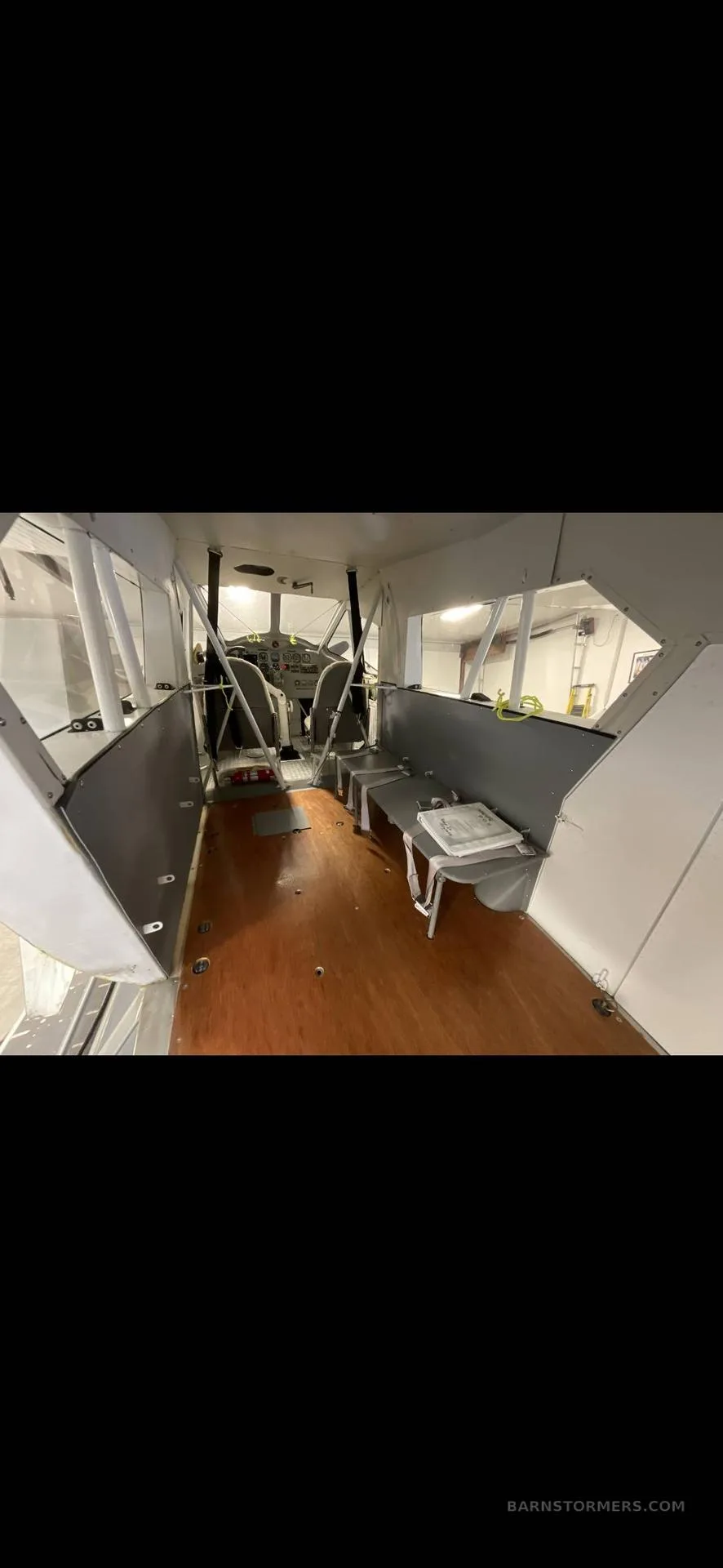 The interior of an aircraft cabin with wooden floorboards, bare walls, and seats visible towards the cockpit.