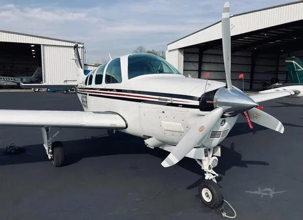 Front 3/4 view of a white Beechcraft Bonanza with black and red stripes on an airport tarmac.