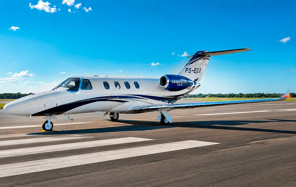 A white and blue Cessna Citation M2 business jet (PS-EGX) parked on an airfield under a clear blue sky.