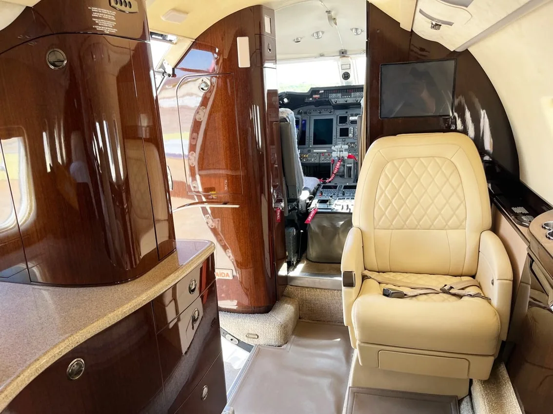 The luxurious cabin interior of a business jet features cream leather seats, dark wood cabinetry, and a view into the cockpit.
