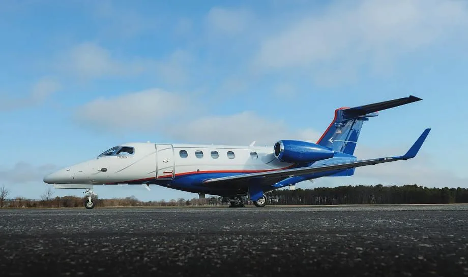 A white and blue Embraer Phenom private jet is parked on an asphalt runway under a partly cloudy sky.