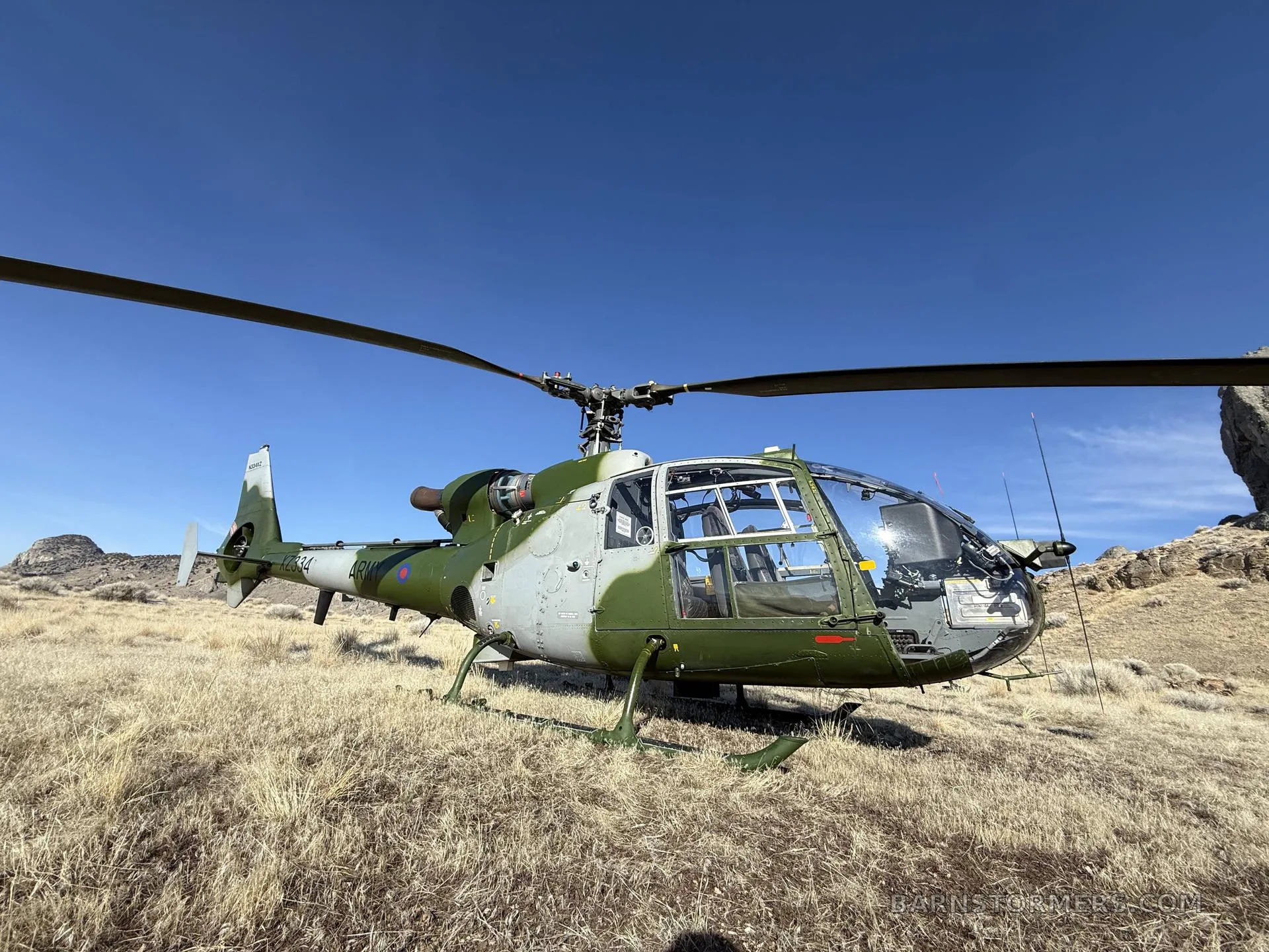A green and grey military Westland Gazelle helicopter is parked on dry, golden grass under a bright blue sky.
