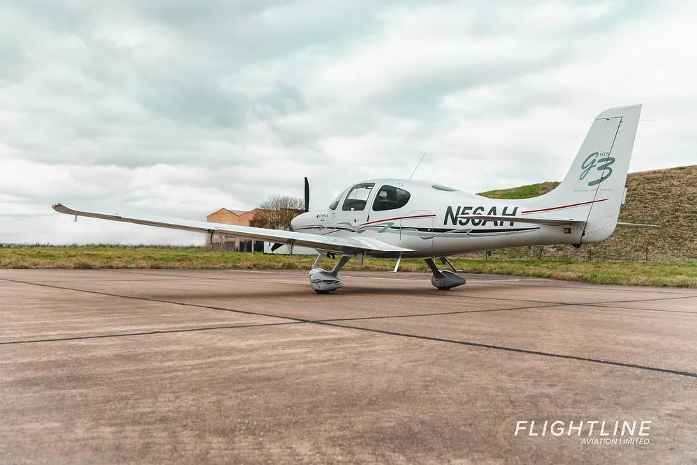 A rear three-quarter view of the white Cirrus SR22 aircraft N56AH on a tarmac with a building in the background.