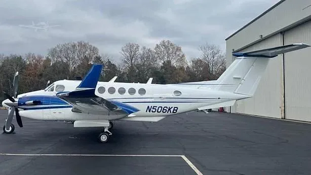 A white and blue twin-turboprop aircraft, N506KB, is parked on an asphalt ramp next to a hangar under an overcast sky.